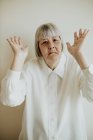 Грустная пожилая женщина в белой блузке демонстрирует несогласие, поднимая руки на светлом фоне, глядя в камеру. — стоковое фото