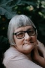 Senior woman with grey hair looking at camera in eyeglasses crossing arms over shoulder among plants with big green leafs — Stock Photo