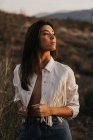 Alluring young charming female brunette wearing denim and white jacket on naked body while standing in hilly terrain — Stock Photo