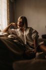 Sensual loira senhora de cabelos brancos camisa sentada no sofá tocando a boca de forma fascinante e olhando para baixo na sala de estar — Fotografia de Stock