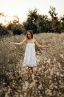 Peaceful young woman in white romantic dress standing and touching tall grass with field flowers and looking at camera — Stock Photo