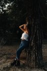 Повна довжина мрійливої жінки в блакитному джинсовому і білому верхньому, спираючись на дерево, дивлячись геть — стокове фото