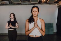 Ruhige schlanke Damen in Aktivkleidung mit Namaste stehen in Bergstellung auf Sportmatten auf Holzboden und meditieren mit geschlossenen Augen in einem geräumigen modernen Fitnessstudio. — Stockfoto