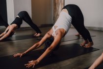Side view of fit barefoot unrecognizable women in sportswear concentrating and doing downward facing dog exercise on sports mats on wooden floor against white walls of spacious hall — Stock Photo