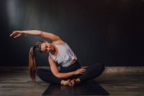 Jeune femme détendue avec les yeux fermés et le bras levé assis en position baddha konasana sur le tapis de sport et pratiquant le yoga dans la salle sombre — Photo de stock