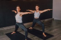 Женщины в спортивной одежде делают воин представляют два упражнения йоги стоя на спортивных ковриках в современной тренировочной комнате — стоковое фото