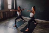 Mujeres en ropa deportiva haciendo guerrero posan dos ejercicios de yoga de pie sobre alfombras deportivas en la sala de entrenamiento moderna - foto de stock