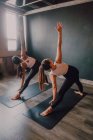 Vista posterior de mujeres irreconocibles haciendo ejercicio de pose de triángulo giratorio de pie sobre alfombras deportivas en la sala de entrenamiento moderna - foto de stock