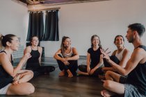 Высокий угол юных женщин и мужчин в спортивной одежде, сидящих в позе лотоса и интересующихся дискуссиями во время отдыха после групповых тренировок в современной студии йоги — стоковое фото