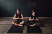 Calm young woman and man in sportswear with eyes closed and legs crossed meditating together while sitting in padmasana position against back wall in contemporary yoga studio — Stock Photo
