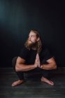 Lächelnder, gut aussehender, flexibler männlicher Athlet in Sportbekleidung mit Namaste, der Yoga-Kakasana-Position auf Holzboden vor schwarzer Wand des modernen Trainingsraums ausübt — Stockfoto