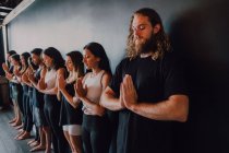 Группа счастливых молодых спортсменов в спортивной одежде с закрытыми глазами, расслабляющими и глубоко дышащими, стоя с намасте в тадасане и медитируя у черной стены в современной студии йоги — стоковое фото