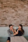 High angle of cheerful man and young woman in casual clothes looking and feeding each other with tasty ice cream while sitting at table on sofa and relaxing together in modern cafe in loft style — Stock Photo