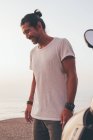 Счастливый взрослый мужчина в футболке, стоящий рядом с мотоциклом на берегу моря на закате — стоковое фото