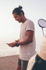 Vue latérale d'un homme sportif moderne positif en t-shirt et short debout à côté du vélo et utilisant un téléphone portable au bord de la mer au coucher du soleil — Photo de stock