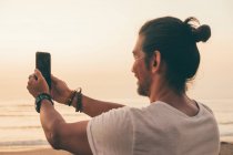 Мужчина фотографирует со смартфоном на пляже — стоковое фото
