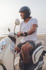 Side view of happy active guy dressed in white t shirt with shorts and black helmet riding scooter in summer evening on beach — Stock Photo