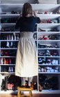 Back view of faceless woman standing on a wooden stool taking beige high heels shoes from shelf of modern white closet — Stock Photo