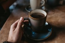 Coltiva mani di donna irriconoscibile seduta a un tavolo di legno che tiene il caffè caldo in una tazza rustica — Foto stock