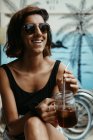 Mujer satisfecha de vacaciones con gafas de sol elegantes mirando hacia otro lado sosteniendo un vaso de cóctel con vista tropical sobre fondo borroso - foto de stock