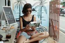 Відпочиваюча жінка в повсякденному одязі і модні сонцезахисні окуляри читають книгу під час відпочинку у відкритому барі — стокове фото