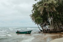 Einsamer Sandstrand mit Holzboot auf welligem Meer in der Nähe grüner tropischer Bäume mit wolkenverhangenem, stürmischem Himmel im Hintergrund — Stockfoto