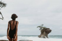 Vista posteriore della donna in costume da bagno nero che cammina sulla costa sabbiosa vuota con oceano tempestoso in nuvoloso sullo sfondo — Foto stock