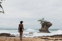 Back view of woman in black swimsuit walking on empty sandy coast with stormy ocean in overcast on background — Stock Photo