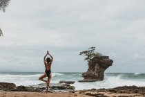 Visão traseira de turista feminino sem rosto em maiô preto equilibrando em uma perna com os braços levantados acima da cabeça na praia vazia na tempestade — Fotografia de Stock