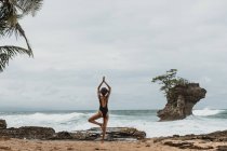 Visão traseira de turista feminino sem rosto em maiô preto equilibrando em uma perna com os braços levantados acima da cabeça na praia vazia na tempestade — Fotografia de Stock