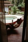 Вид сбоку на мужчину в купальном костюме, сидящего на гамаке и читающего книгу у бассейна отеля-курорта — стоковое фото
