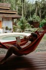 Vista laterale del viaggiatore maschio in costume da bagno agghiacciante su amaca e libro di lettura a bordo piscina dell'hotel resort — Foto stock