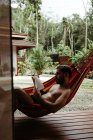 Вид сбоку на мужчину в купальном костюме, сидящего на гамаке и читающего книгу у бассейна отеля-курорта — стоковое фото
