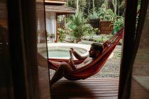 Вид збоку чоловічого мандрівника в купальнику, який охолоджує гамак і читає книгу в басейні курортного готелю — стокове фото