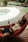 Dall'alto vista laterale del viaggiatore maschio in costume da bagno che si rilassa su amaca e libro di lettura a bordo piscina dell'hotel resort — Foto stock