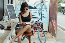 Отдыхающая женщина в повседневной одежде и модных солнцезащитных очках читает книгу во время освежения в баре на открытом воздухе — стоковое фото