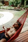 Сверху вид на мужчину-путешественника в купальнике охлаждения на гамаке и чтение книги у бассейна курортного отеля — стоковое фото