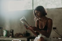 Заинтересованная молодая женщина в бюстгальтере и коротком наслаждаясь чтением книги, сидя со скрещенными ногами на прилавке на кухне — стоковое фото