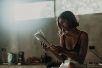 Interessante Frau in BH und kurzer Hose, die es genießt, ein Buch zu lesen, während sie mit gekreuzten Beinen auf der Marmorarbeitsplatte in der Küche sitzt — Stockfoto