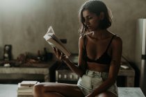 Interessante junge Frau im BH und kurzer Freude am Lesen eines Buches, während sie in der Küche auf einer Marmorarbeitsplatte sitzt — Stockfoto