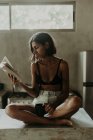 Mujer joven interesada en sujetador y corto disfrutando de la lectura de un libro mientras está sentado en la encimera de mármol en la cocina - foto de stock