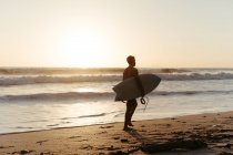 Вид ззаду на силует людини, що тримає дошку для серфінгу під час прогулянки вздовж піщаного узбережжя влітку під час заходу сонця — стокове фото