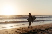 Rückansicht der Silhouette eines Mannes mit Surfbrett, während er im Sommer bei Sonnenuntergang entlang der sandigen Küste läuft — Stockfoto