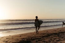 Vue arrière de la silhouette de l'homme tenant la planche de surf tout en marchant le long du littoral sablonneux en été pendant le coucher du soleil — Photo de stock
