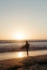Side view of man silhouette holding surfboard while walking along sandy seashore in summertime during sunset — Stock Photo