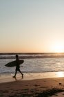 Side view of man silhouette holding surfboard while walking along sandy seashore in summertime during sunset — Stock Photo