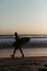 Вид збоку силует людини тримає дошку для серфінгу під час прогулянки вздовж піщаного узбережжя в літній час під час заходу сонця — стокове фото