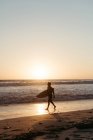 Vista laterale della silhouette dell'uomo che tiene la tavola da surf mentre cammina lungo la spiaggia sabbiosa in estate durante il tramonto — Foto stock