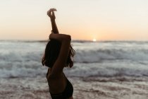Seitenansicht einer nicht wiedererkennbaren Frau im Bikini, die die Hände hebt und im sommerlichen Sonnenuntergang das Meer betrachtet — Stockfoto