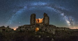 Desde abajo vista trasera de un turista anónimo con linterna explorando el viejo castillo en ruinas bajo la Vía Láctea en la noche estrellada - foto de stock
